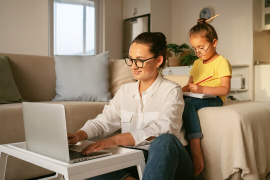 Young Smiling Woman Mother Freelancer Working Remotely At Home With Small Child, Mom Using Laptop While Daughter Sitting On Sofa And Drawing. Remote Job And Family Concept