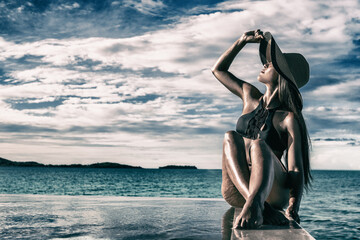 Elegant black swimsuit model woman high end fashion with sun hat on infinity poolside luxury resort...