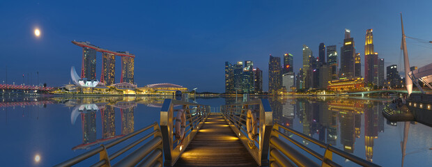 Vibrant panoramic view of Singapore skyline from a jetty at blue hour with nice reflection and full moon rising