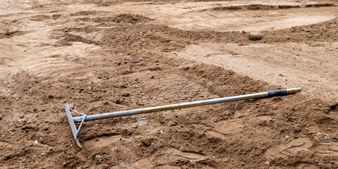 A rake with metal handle, used for leveling the earth and dirt, lays on the brown soil at a house...