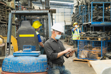 Men work together, wear safety facemask, using tablet in medium shot. Caucasian engineer men using tablet while Asian men working behind him in factory-warehouse