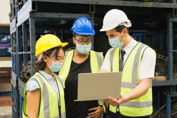 Men and woman work together, wear safety facemask and using laptop. Black women looking at screen on laptop while Caucasian engineer men discuss with Asian engineer women in factory-warehouse