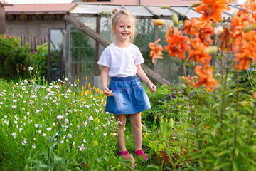 little girl stands in meadow of flowers and laughs, against background of greenhouse. Carefree childhood, happy child.