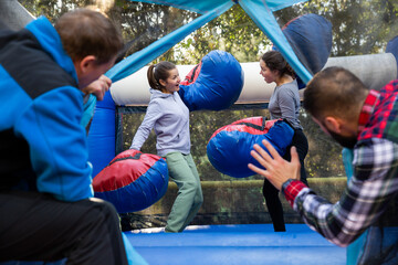 Obraz premium Friends having fun in outdoor amusement park, two men watching women in big boxing gloves boxing on inflatable ring