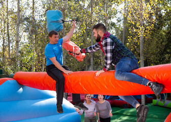 Laughing man sitting on inflatable log and fighting by pillows with his friend on adults bouncy...