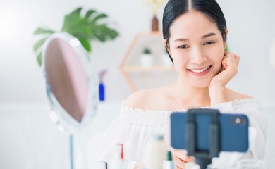 Beautiful asian woman blogger is showing how to make up and use cosmetics. In front of the mirror and smartphone recording vlog video live streaming at home.Skincare for healthy face concept.