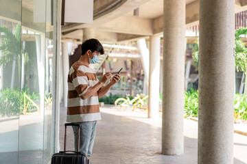 Travel. A man wearing glasses with a striped shirt and mask is using mobile phone during travel with a suitcase placed on his right hand