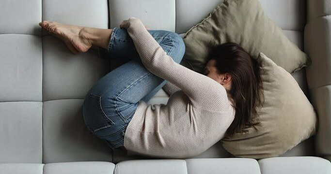 Woman lying on couch curled up suffers from menstrual cramps, above view. Unplanned pregnancy and abortion, try to sleep hiding from life troubles, drugs alcohol addict, break up and divorce concept