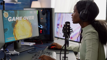 African streamer woman playing on powerful RGB computer shooter video game losing tournament, talking with multiple players into headphones. Pro video gamer using professional streaming equipment