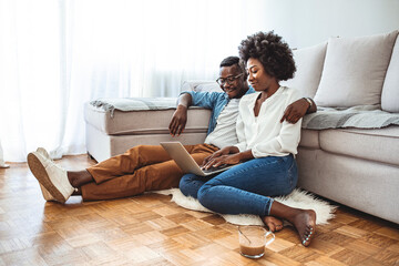 Fototapeta premium Young black couple sit on the floor using laptop, side view. People, leisure, mutliethnic relationship concept. Lovely mixed race couple spend free time at home, dressed casually, embrace