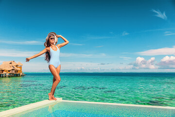 Travel vacation tourist woman relaxing at infinity pool in French Polynesia. Happy woman in swimsuit and sun hat over blue ocean at luxury resort. Elegant lady enjoying holiday lifestyle.