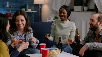 Multi-cultural friends laughing while sharing lifestyle advice sitting on couch late at night in living room. Multi-ethnic people hanging out, drinking beer enjoying together during wekeend party.