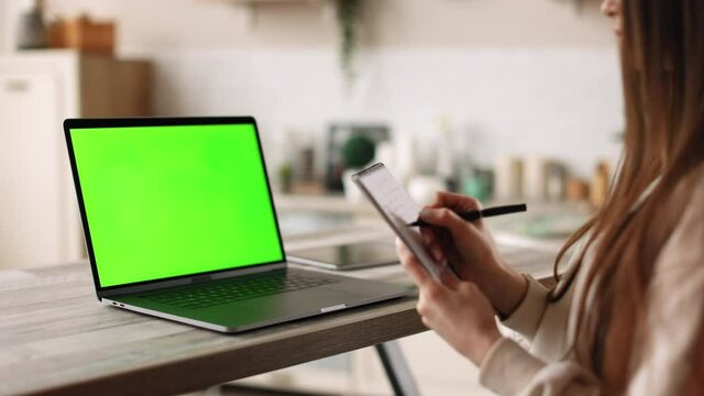 A woman is sitting in her home office. She is making notes in her notebook. On the table is a laptop with a green screen. Chromakey. 4K