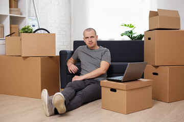 moving day concept - man sitting on floor with laptop and cardboard boxes in room after moving day