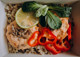 white and brown rice with red fish meat, chopped paprika, seeds and a slice of lime. food in a white paper container closeup. delivery of healthy food for weight loss top view.