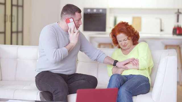 Worried Caucasian Man Talking On The Phone Calling 911 As Ill Woman With Severe Stomach Pain Lying On Couch In Living Room. Portrait Of Loving Husband Taking Care Of Sick Wife At Home Calling