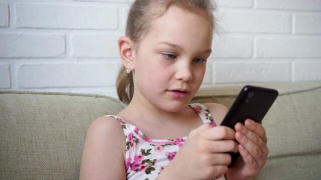 Girl playing mobile game on smartphone on sofa. Preschooler playing mobile phone. Kid using phone for gaming. Child playing video game at home