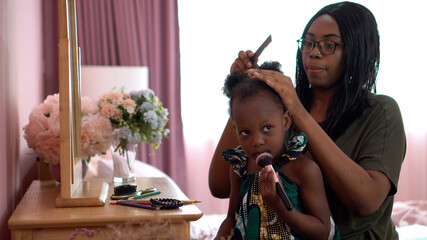 african american mother combing  her little daughter hair sitting near mirror on dressing table in the children room at home. black mom and girl at bedroom in morning time together - Powered by Adobe