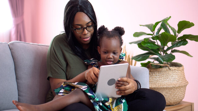 african american mother using tablet with her little daughter on couch at home . black girl lying on her mom learning online at home on sofa together . quarantine Home School