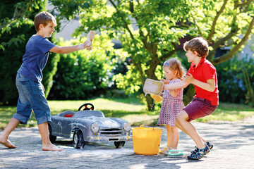 Three happy children washing big old toy car in summer garden, outdoors. Two boys and little toddler girl cleaning car with soap and water, having fun with splashing and playing with sponge.