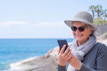 Attractive and fashionable senior woman smiles on beach vacation looking at smart phone. Joyful lifestyle concept. Sunny day, blue sky