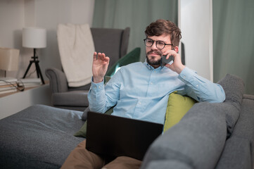 While working from home, a bearded young guy makes a work call, he sits on the couch with a laptop and a smartphone