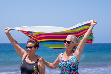 Two mature women enjoying the beach together holding towels in the wind, active retired seniors and vacation concept. Horizon over water