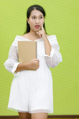 Image is portrait. Asian smiling plump woman wearing white dress. Lady asia standing finger poked on the cheek and holding hand book in green background.