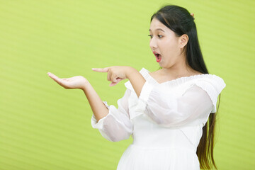 Image is portrait. Asian smiling plump woman wearing white dress. Lady asia standing use her index finger on  hand and shocked expression in green background.