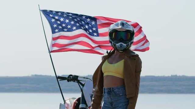 An American motocross racer stands in front of the waving US flag and turns his head towards the video camera.