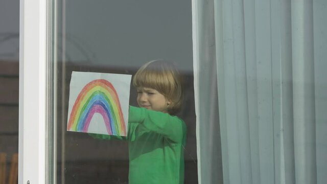 Child with rainbow drawing at window, home self isolation, hope message