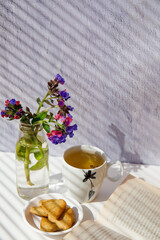 Beautiful pink and purple wood lungwort (Pulmonaria officinalis) in glass vase with cup of green tea and cookies on white background with striped shadows. Spring flowers. Vertical, copy space