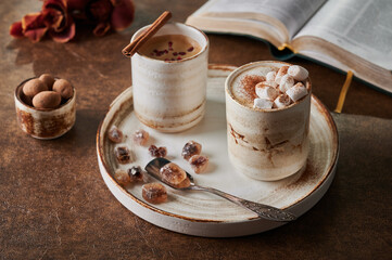 Two cups of coffee with foam and marshmallow and sublimated raspberries with cinnamon on light plate with caramelized sugar and spoon. Nearby is an open book, flowers and candy. Selective focus