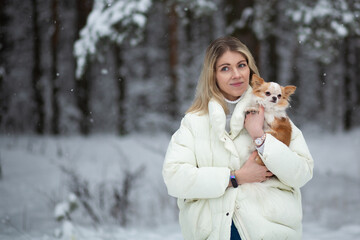 Blonde young female holding ginger and white chihuahua in her hands. Snow and trees on the background