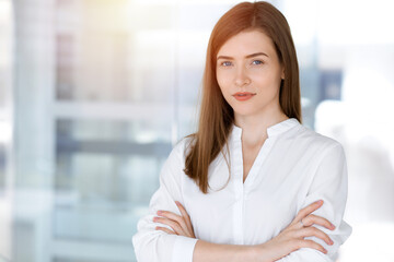 Beautiful female specialist with arms crossed standing in sunny office and smiling charmingly. Business people concept