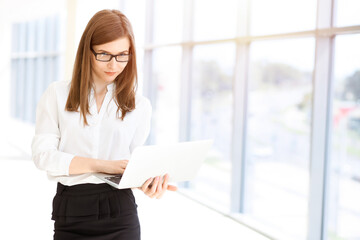 Beautiful female specialist with laptop computer standing in sunny office and smiling charmingly. Business people concept
