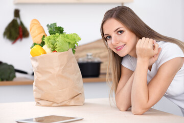 Young brunette woman smiling while sitting at the table near paper bag full of vegetables and fruits. Concept of food buying and kitchen lifetime