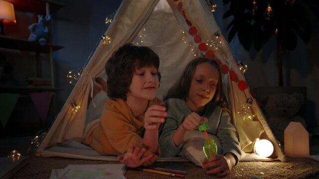Kids in cute decorative makeshift tent blowing bubbless at home in evening. Cheerful girl and boy lying on floor while spending good time. Concept of leisure and careless childhood.