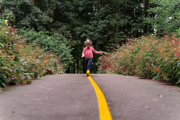 Obraz premium little girl in yellow rubber boots walks along the road in the park on a summer day