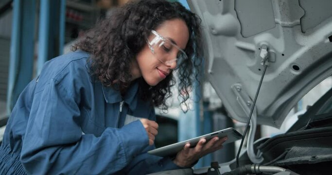 Female mechanic uses tablet computer with diagnostics software checking car engine. Specialist inspecting the car in order to find broken components inside the engine bay. Car Service.