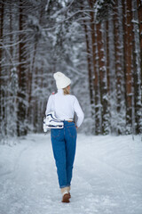 Young blond beautiful female with white ice skates in her hand in winter snowy forest.