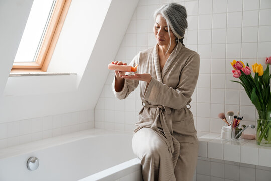 Mature grey woman applying cream while sitting in bathroom at home