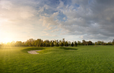 Golf course with lawn. Evening landscape in the park. Blue sky with clouds and bright sunlight with rays at sunset.