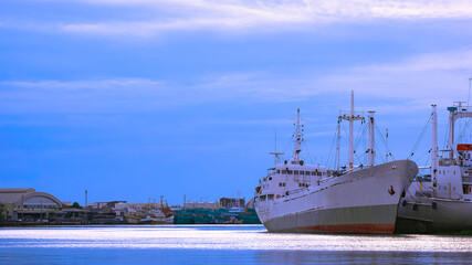 Selective focus at large fishing boats with many vessel moored at harbor in industrial community along the coast against blue sky in evening time