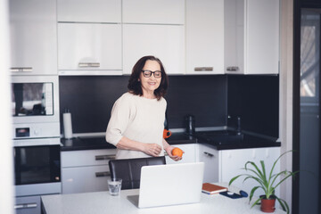 Fototapeta na wymiar Beautiful happy mature elderly woman with orange in her hands standing at home in the kitchen in front of laptop screen, healthy eating and active retirees concept