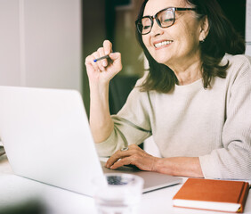 Beautiful mature happy woman 60 years in glasses working remotely online at home on laptop,...