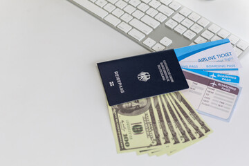 Closeup shot of one hundred dollar US banknotes currency heap covered by blue passport with air tickets boarding pass on working desk with computer screen monitor keyboard mouse and succulent pot
