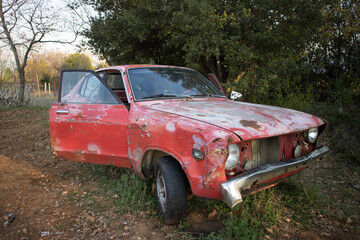 An old car with faded red colour and open doors. At a vehicle graveyard.