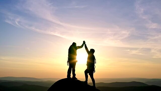 Silhouettes of a man and a woman giving high fives on top of a rocky mountain range at sunset.
