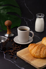A white coffee cup was placed next to a milk bottle and a croissant on a cutting board, coffee beans and grinders on a marble floor.